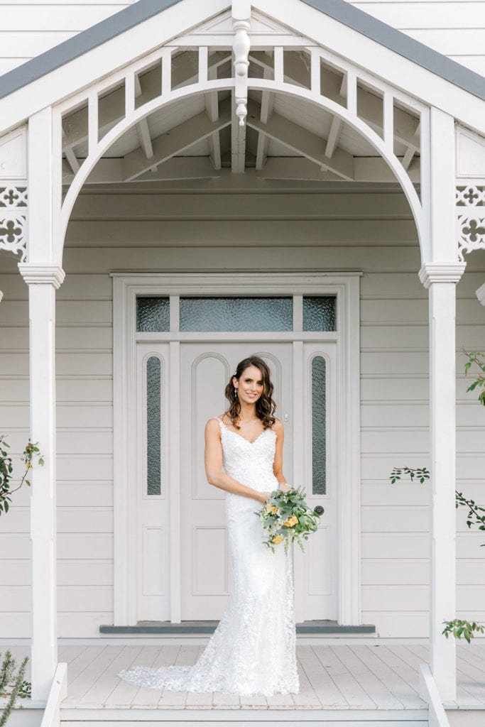 Model wearing Vinka Design Camille Wedding Dress, a Timeless Fitted Wedding Gown on the veranda of a beautiful New Zealand home holding a bouquet