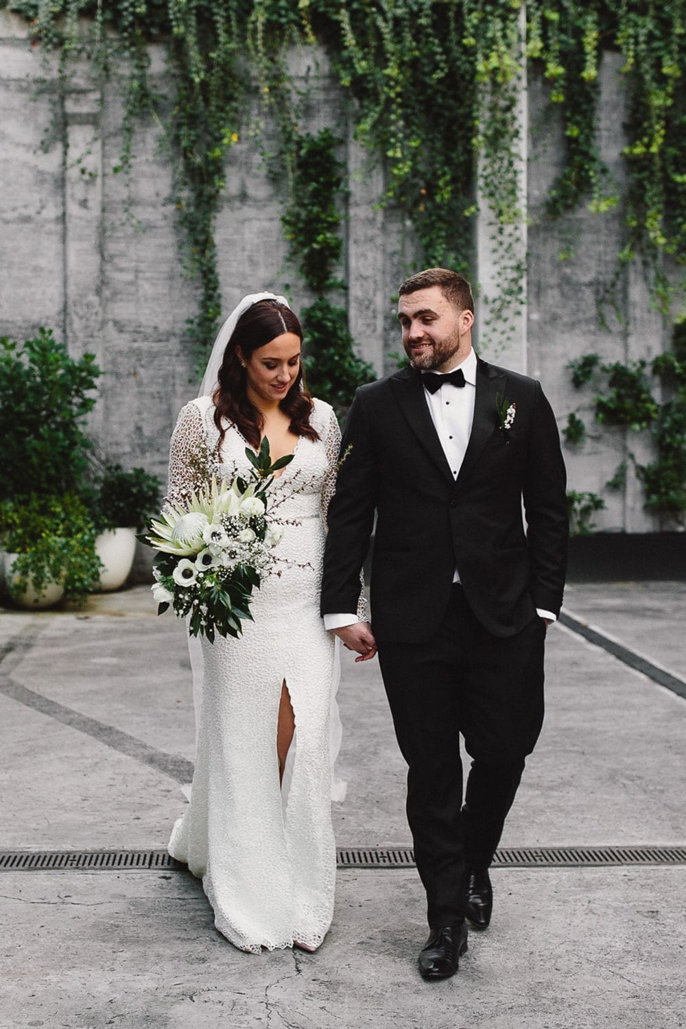 Vinka Design Features Real Weddings - bride in custom made gown fitted with a deep V neckline, front split and long lace sleeves. With groom walking outside in concrete courtyard