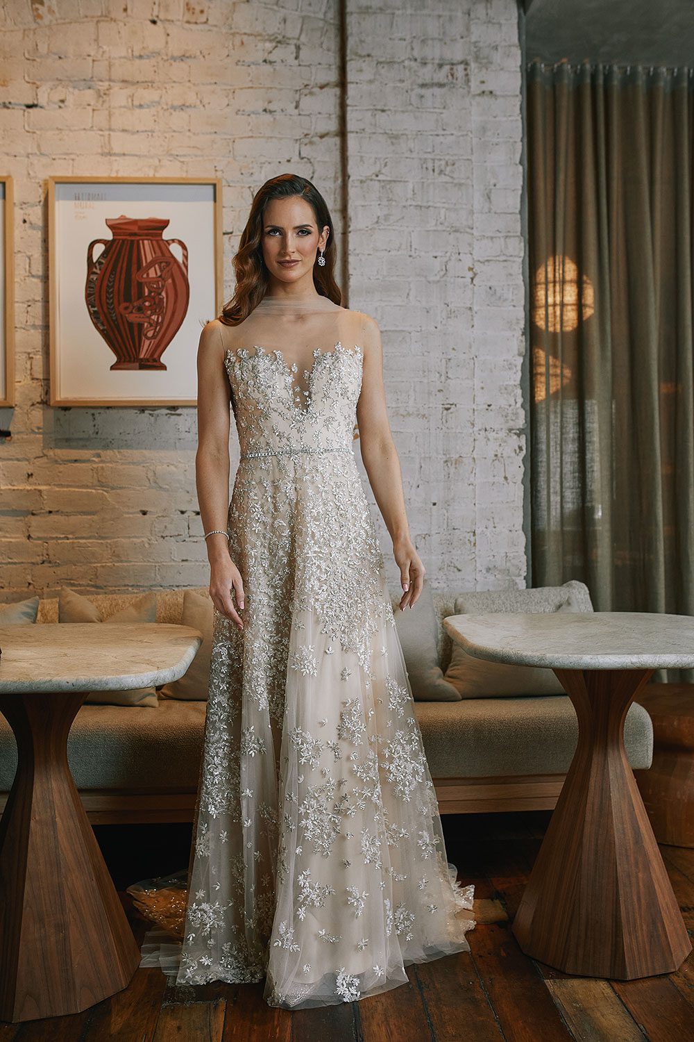 Dita Wedding Dress from Vinka Design. Vintage glamour and modern decadence encapsulates the aura of this gorgeous gown. Featuring richly beaded, botanical tulle embroidered with a dazzling display of silver sequins that glimmer with movement. The high neck, tulle bodice is cut with a plunging neckline, the low cut, scoop back is elegantly adorned with illusion tulle and strategically appliquéd lace on a champagne and blush satin and organza base. Full length view of model in a hotel wearing stunning vintage glamour dress with A-line skirt and cascading train.