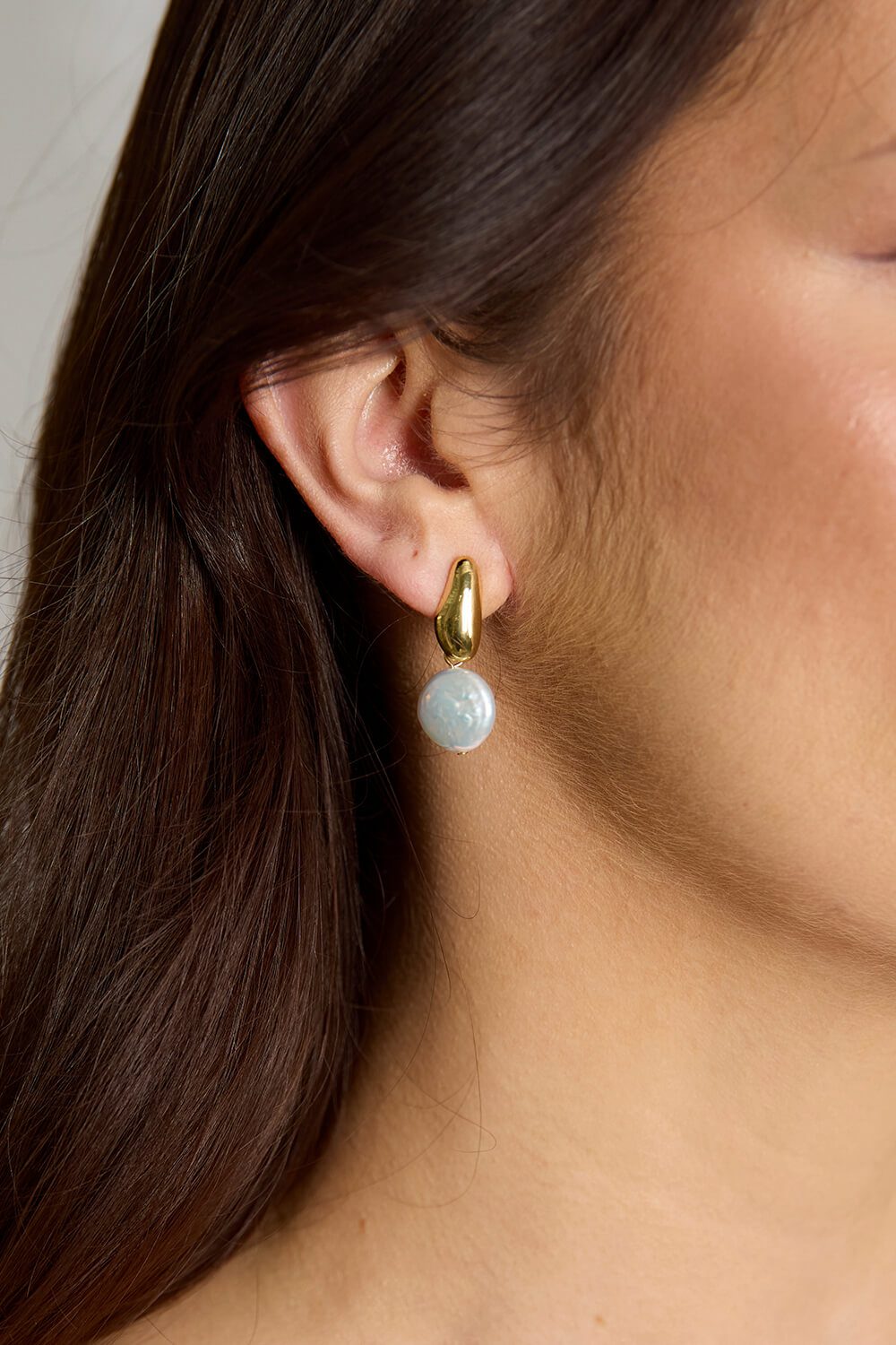 Close-up of the Tina earrings featuring a sculptural gold-plated stud and a luminous white baroque-style pearl drop, worn by a model with dark hair.
